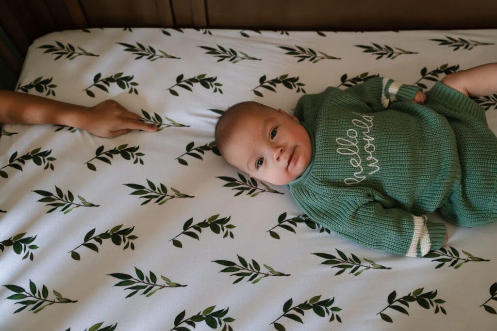 Baby lying on a bed with a green outfit while a sibling reaches toward them during a candid in-home family photography session.