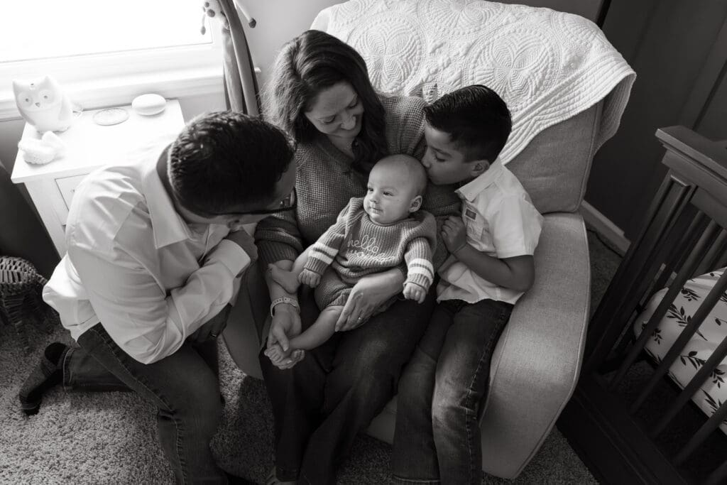 Parents and sibling gathered around a baby sitting on their mother's lap in a cozy chair during an in-home lifestyle family photography session.