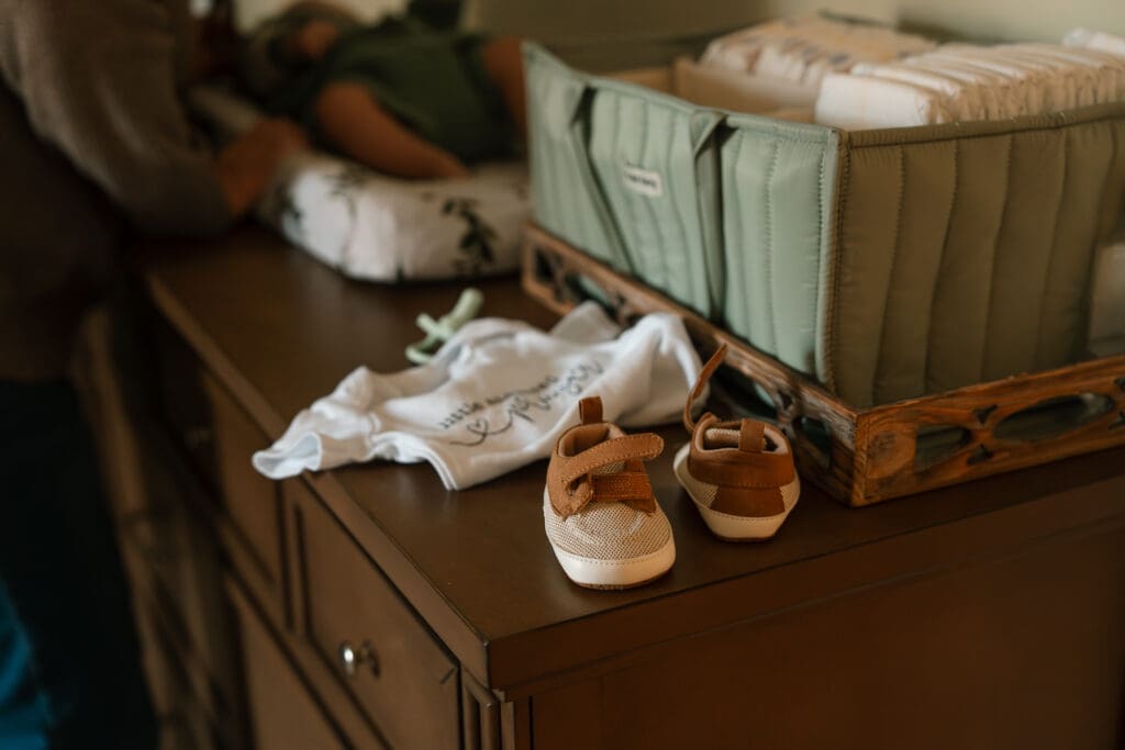 Newborn baby shoes and clothing laid out on a dresser as part of a cozy in-home family photography session.