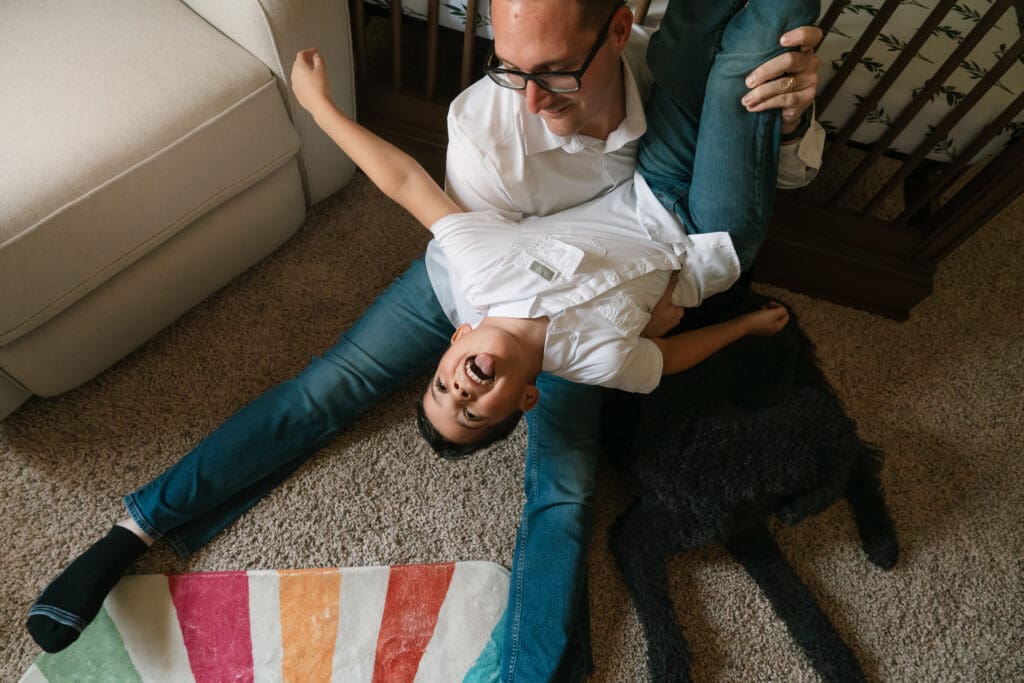 Father sitting on the living room floor with his child playfully draped across his legs during a candid in-home family photo session