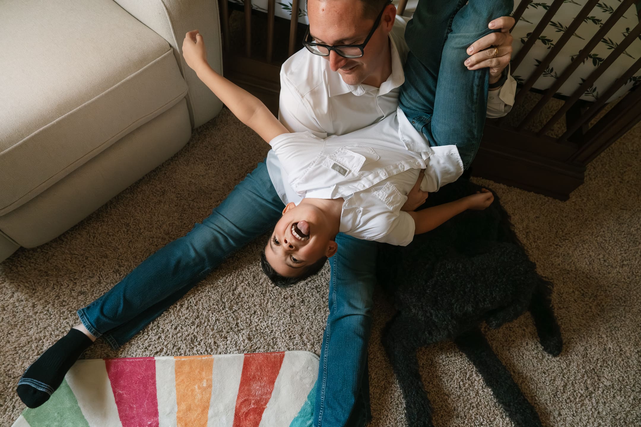 Father sitting on the living room floor with his child playfully draped across his legs during a candid in-home family photo session
