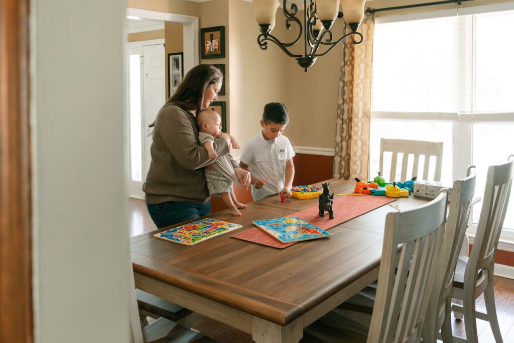Mother holding baby beside a dining table while an older sibling stands nearby during a candid in-home family photography session.