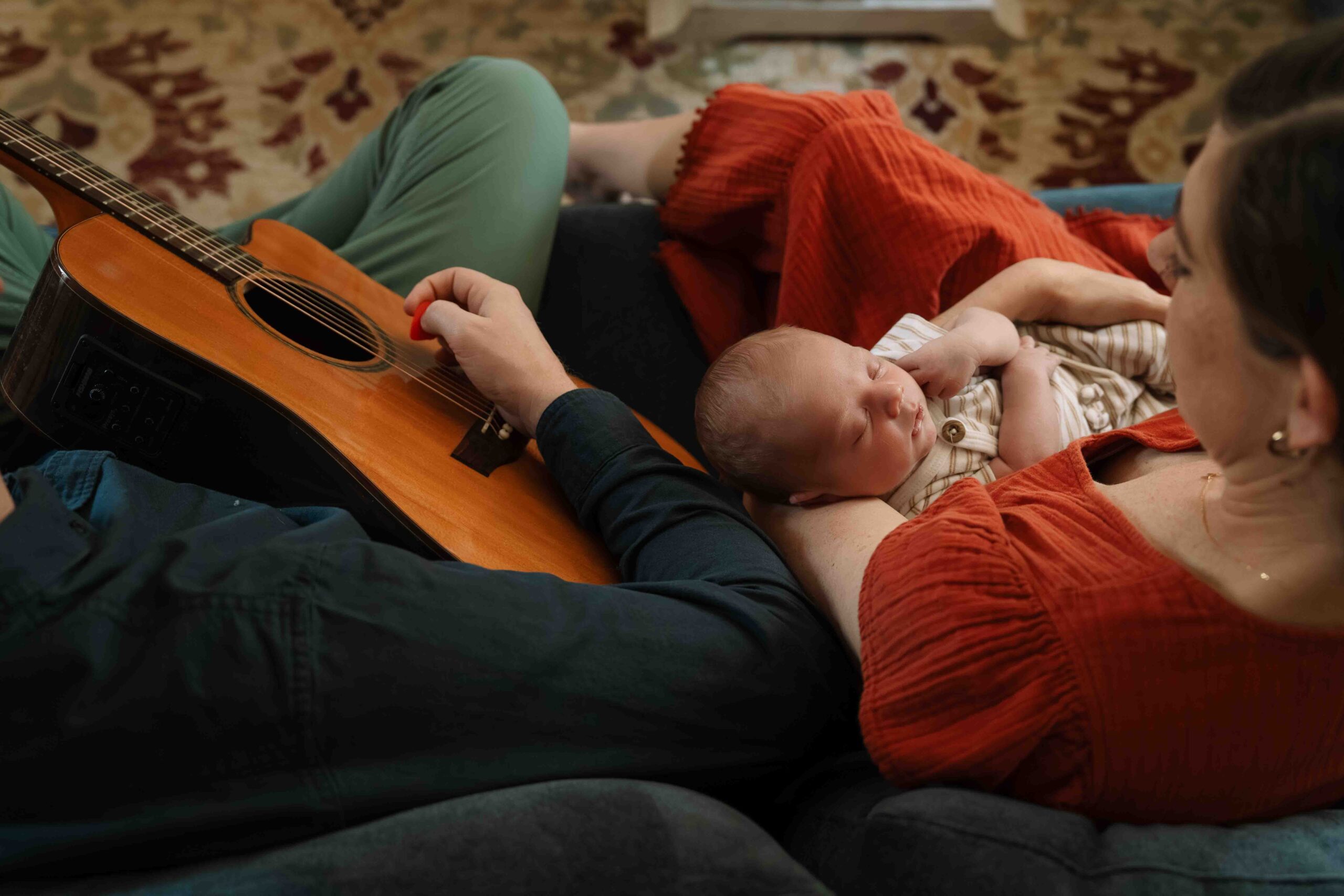 picture of mom and newborn on couch while dad plays guitar