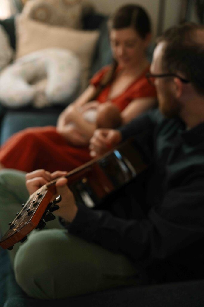 picture of mom and newborn on couch while dad plays guitar
