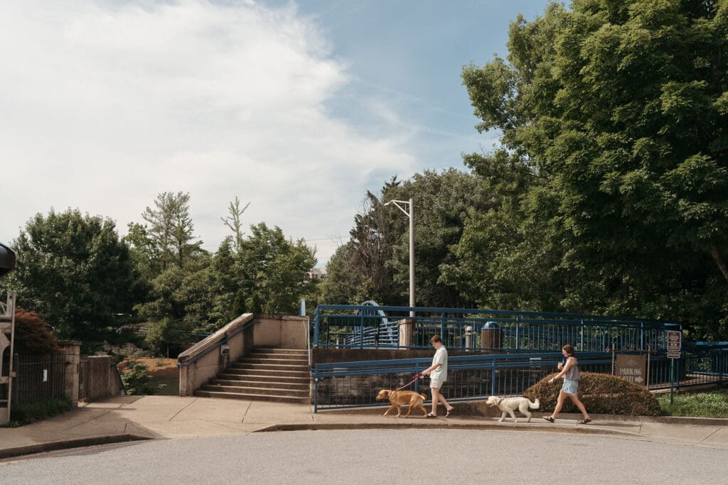 A couple walks their two dogs along a quiet downtown sidewalk near a blue pedestrian bridge on a sunny day.