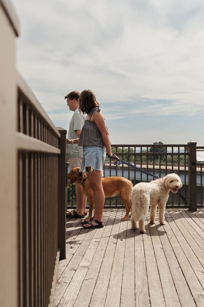 A couple pauses with their two dogs on a scenic boardwalk overlooking the riverfront in downtown Chattanooga on a sunny day.