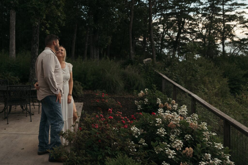 A couple stands close together on a garden overlook at dusk, surrounded by blooming flowers and a wooded mountain view.