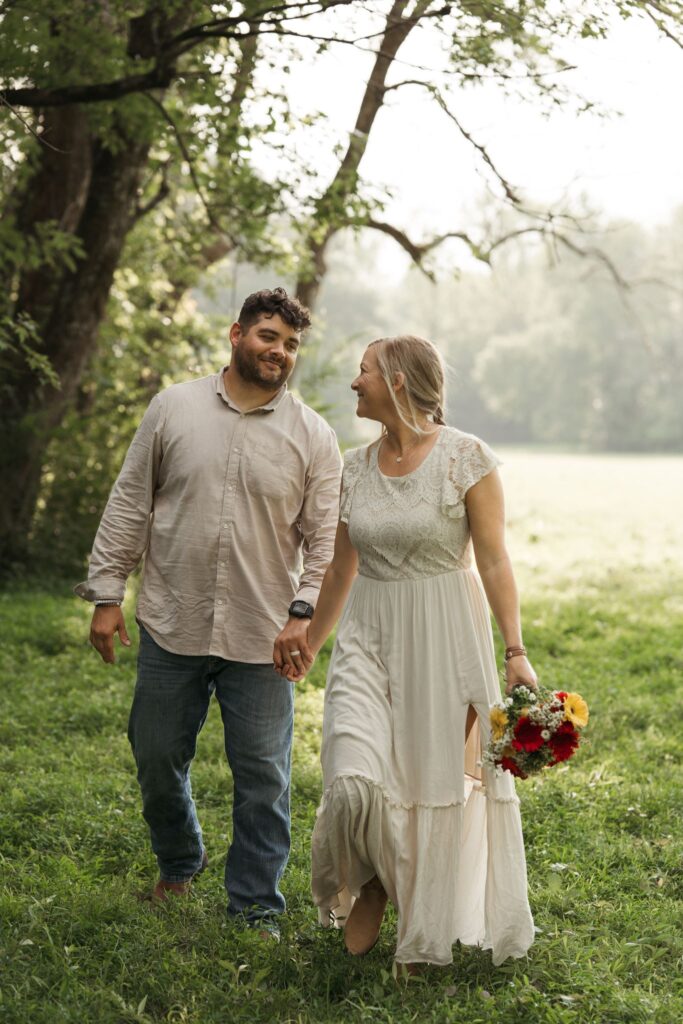 A bride in a flowy dress walks hand in hand with her partner through a misty green field, holding a colorful wildflower bouquet.