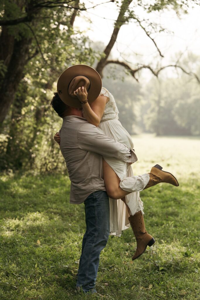 A groom lifts his bride into the air for a joyful embrace in a sunlit, tree-lined field during their elopement.