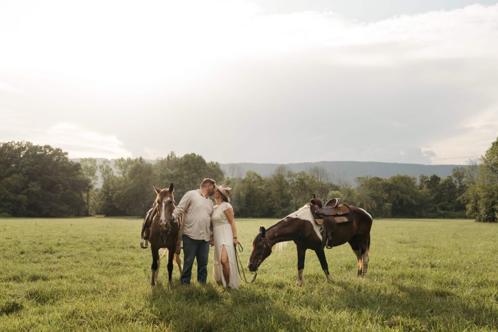 A couple shares a quiet embrace between two horses in an open field with mountains and trees stretching into the distance."