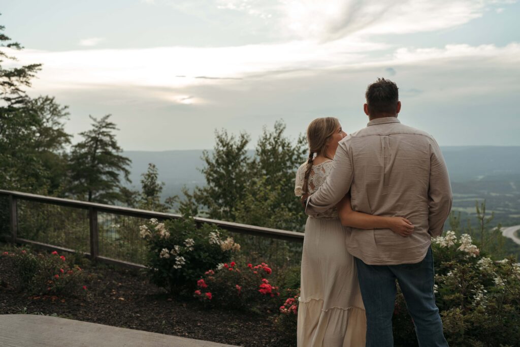 A couple stands arm in arm on a mountain overlook, taking in a sweeping sunset view over the valley below.