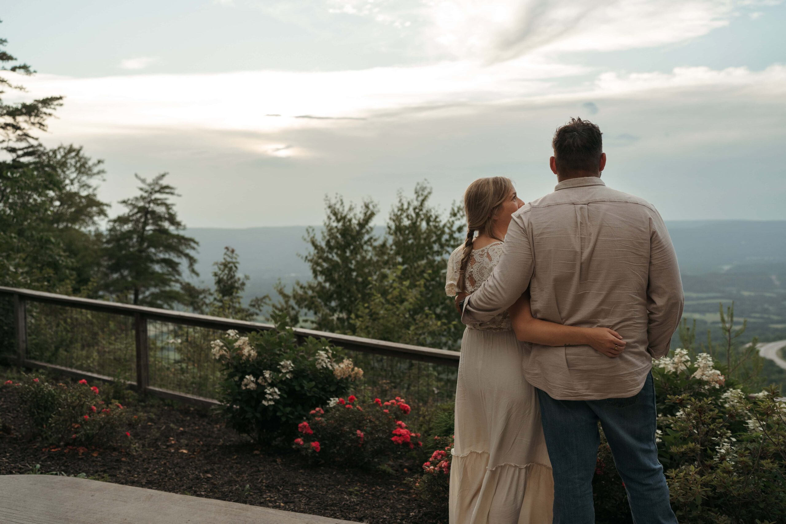 A couple stands arm in arm on a mountain overlook, taking in a sweeping sunset view over the valley below.