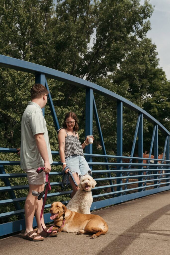 A couple stands smiling on a blue pedestrian bridge with their two dogs resting and sitting beside them.
