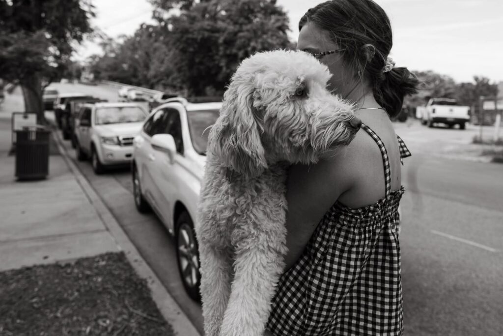 A black-and-white photo of a woman carrying her curly-haired dog over her shoulder on a city sidewalk.