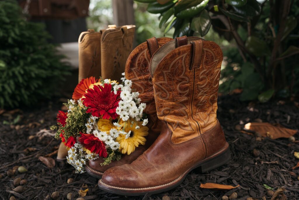 Two pairs of brown leather cowboy boots stand beside a bright bouquet of red and white wildflowers on a wooded forest floor.