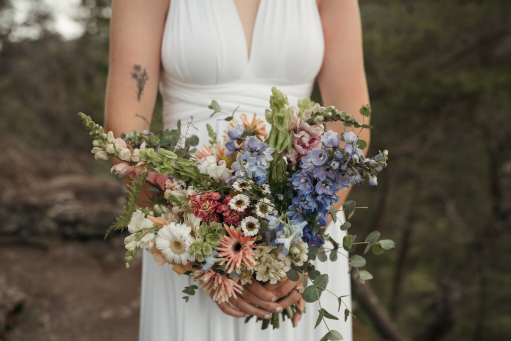 Bride holding a wildflower bouquet with blue delphinium, pink snapdragons, and daisies in a white halter wedding dress