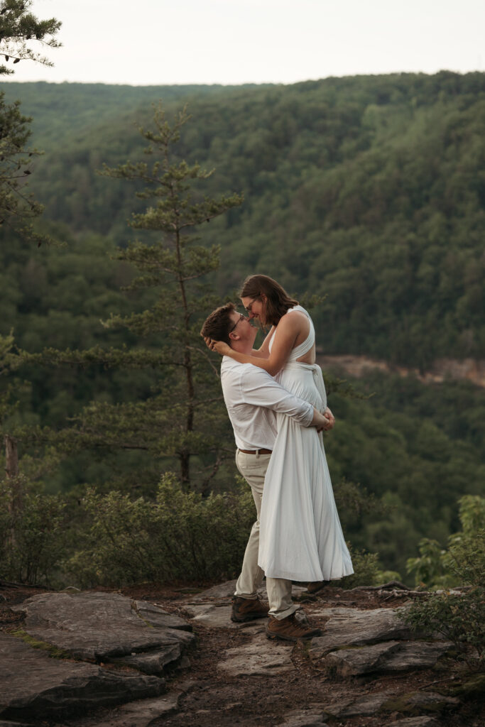 Groom lifting his bride in a hug on a rocky cliffside overlooking a forested valley