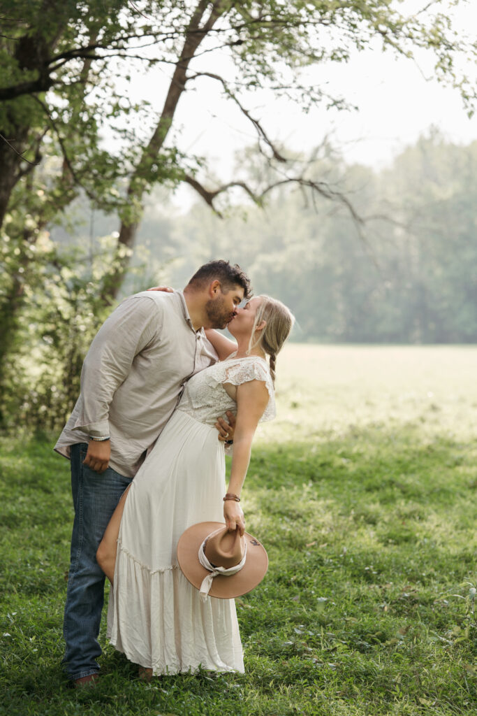 Groom kissing his bride under a tree in a sunlit field, she's holding her hat during their Tennessee elopement