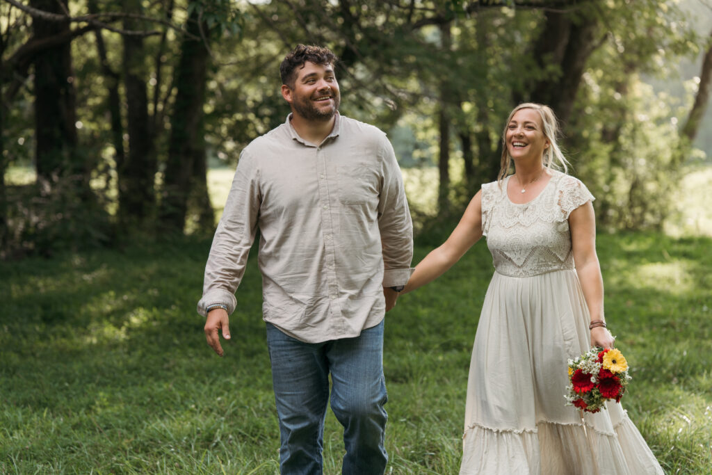 Laughing couple holding hands and walking through a wooded field, bride carrying a bouquet of red and yellow flowers