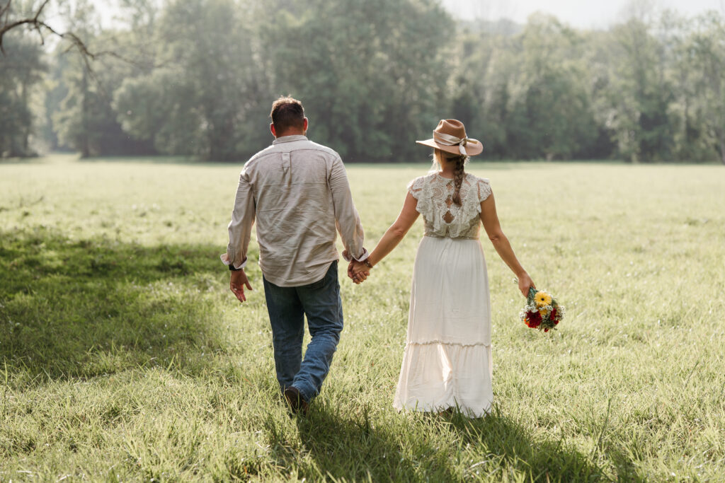 Couple walking hand in hand through an open field, bride wearing a wide-brim hat and carrying a bouquet