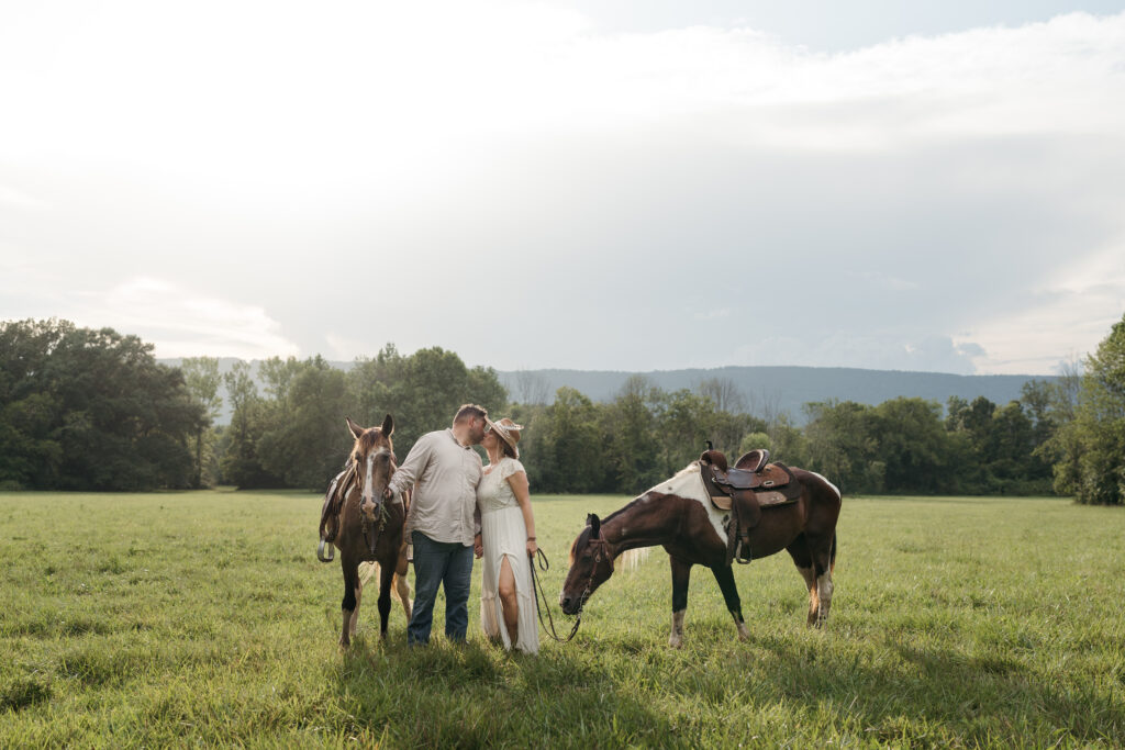 Couple in western wear sharing a kiss beside two horses in a Tennessee mountain field during their elopement